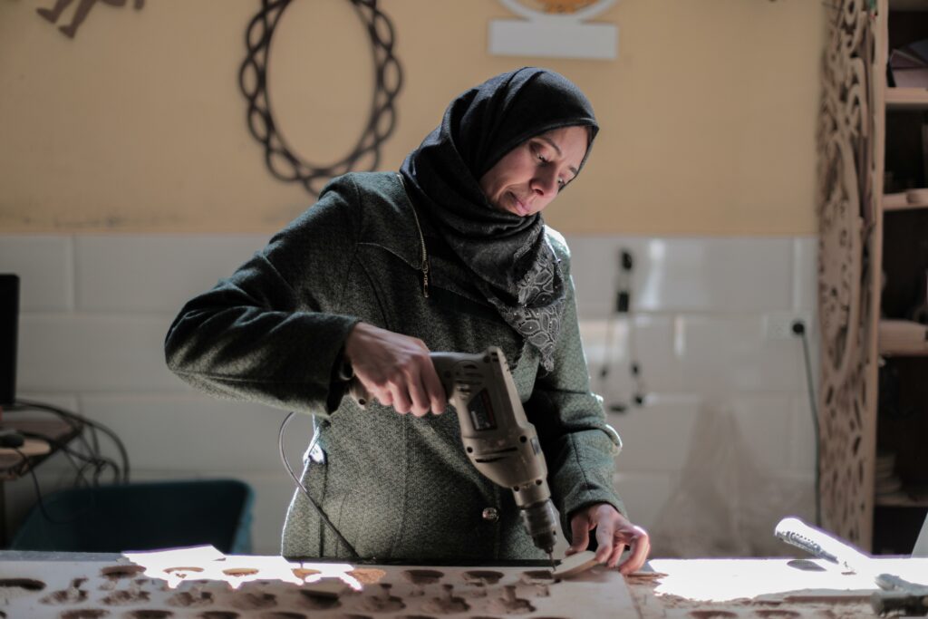 Woman wearing head-covering confidently uses a powered drill to drill a hole in a piece of wood.